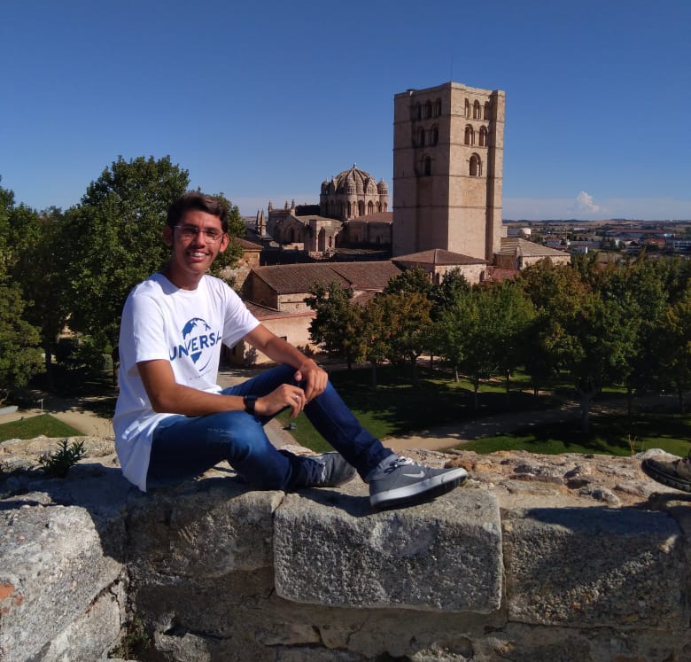 garoto sentado em pedra com monumento ao fundo e sorrindo para a foto